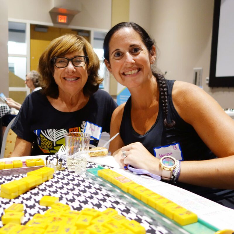 Two women smile while sitting at a game table during an event at the J.