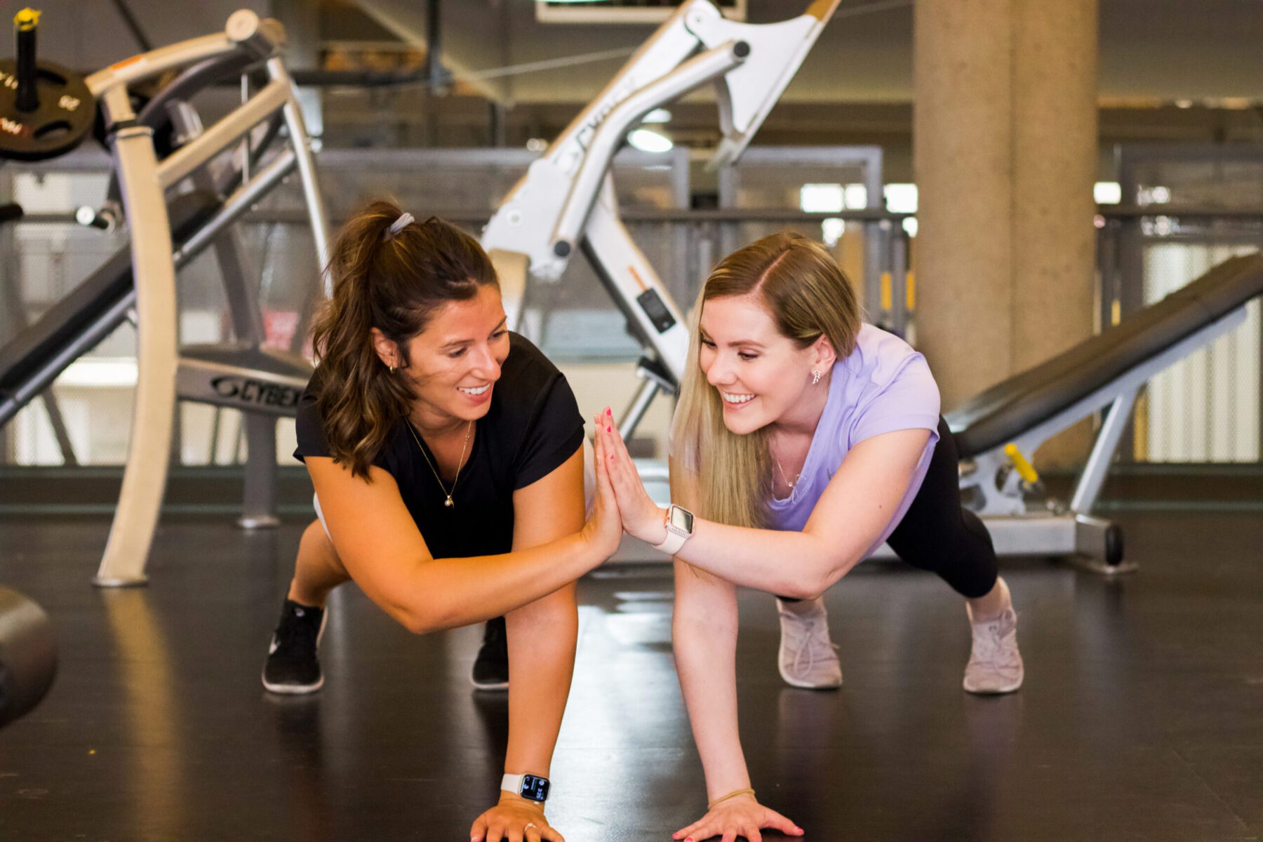 Two women high-five while in a plank position on the floor of a gym.